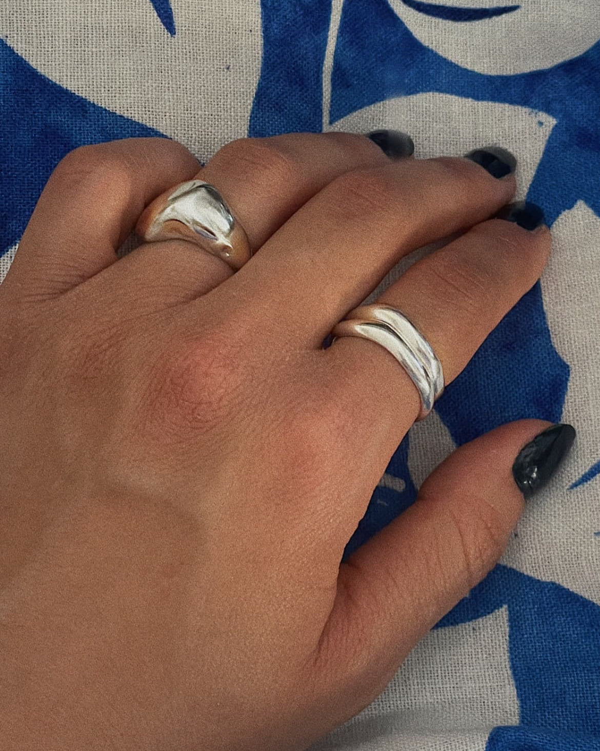 Hand wearing two silver rings from the brand All Rivers with a blue and white floral background