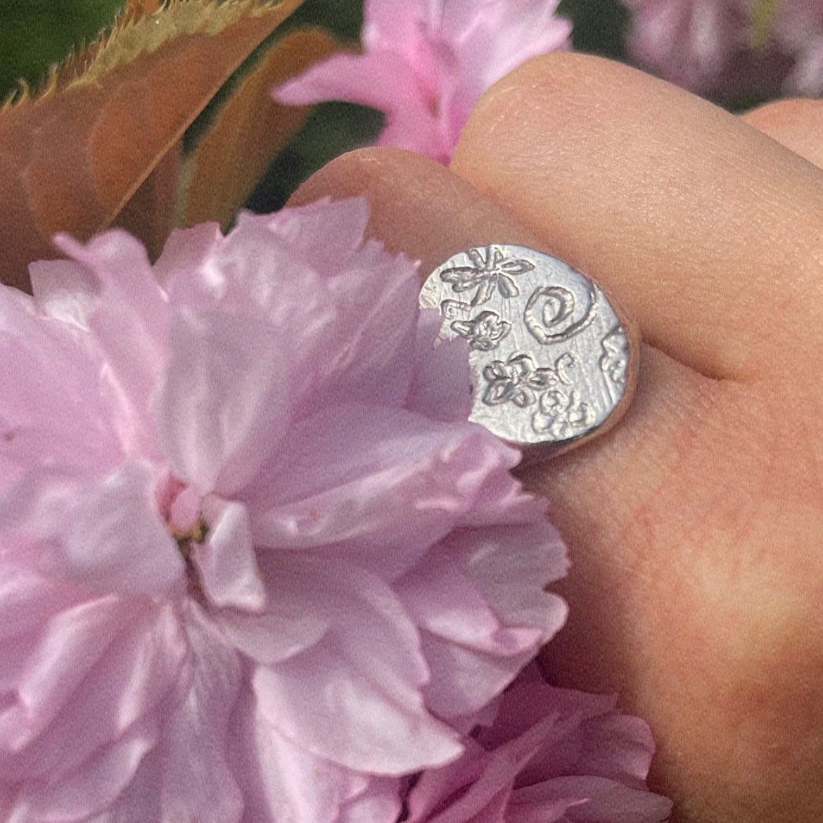 Hand wearing a silver ring by All Rivers, with a floral design, holding pink flowers against a blurred green background