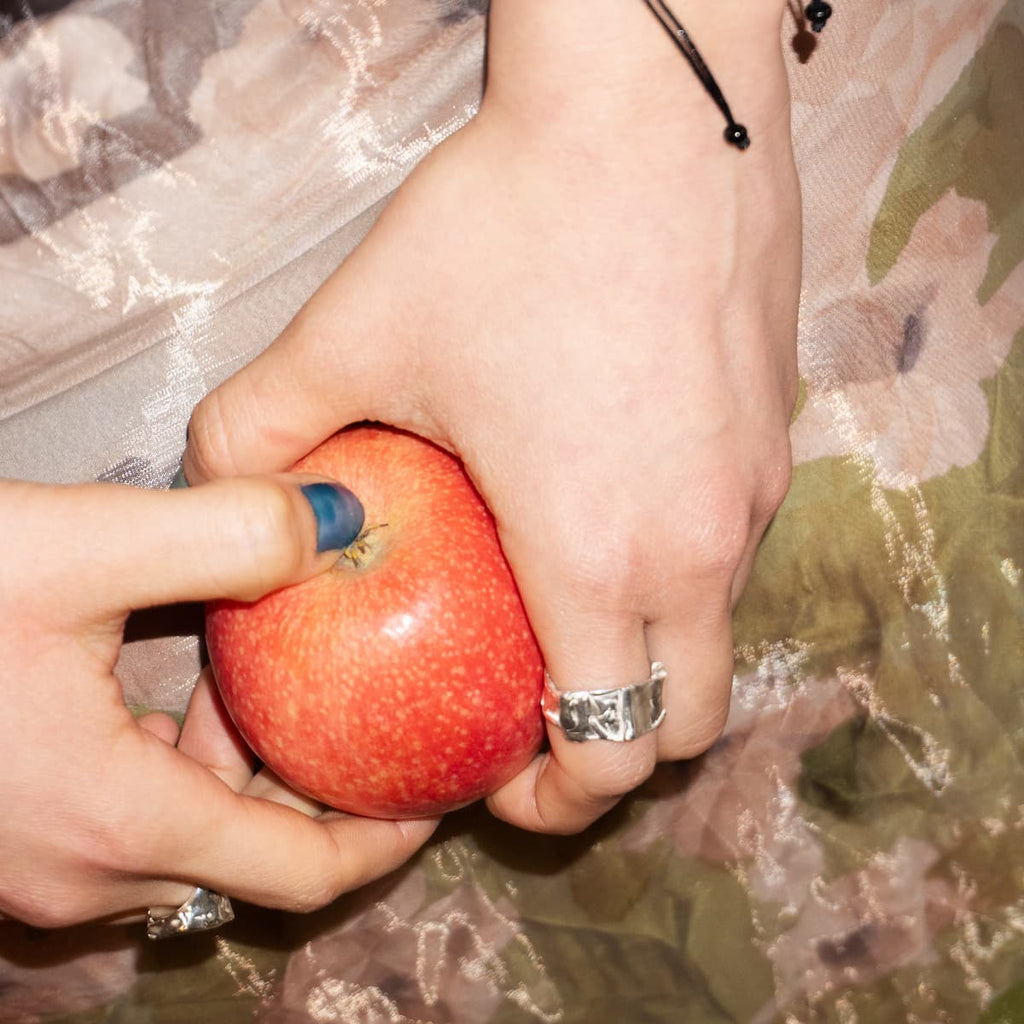 Two hands holding a red apple against a floral fabric background All Rivers Jewelry Yosun Ring from Berlin 