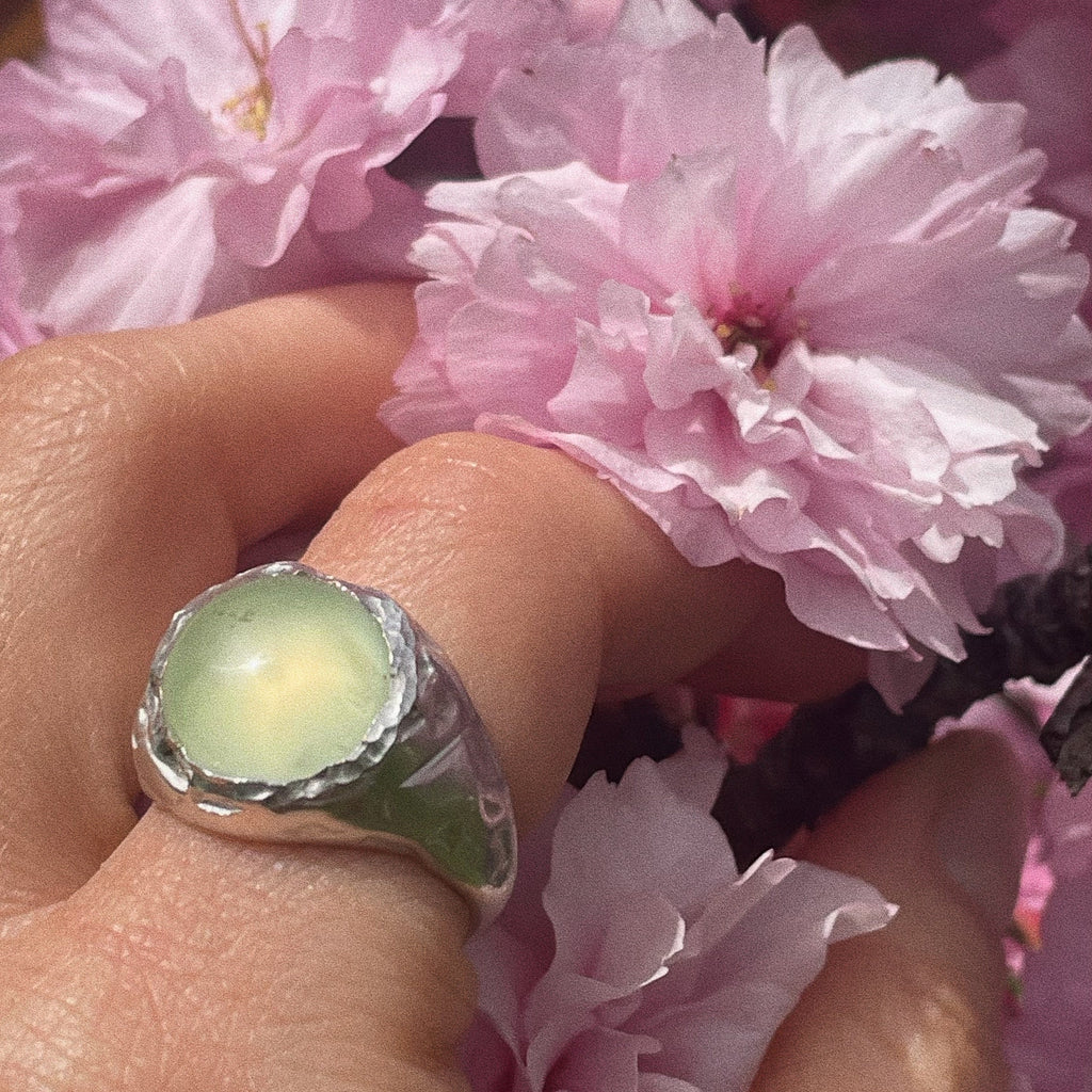 Close-up of a hand wearing a ring with a green stone, surrounded by pink flowers.