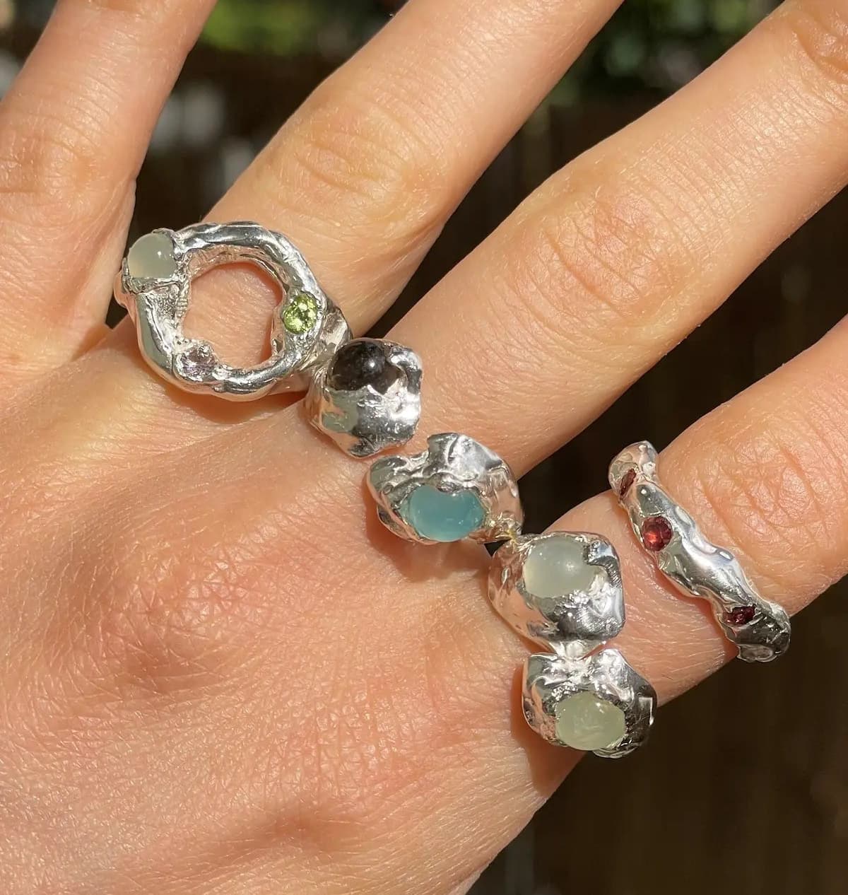 Hand wearing multiple silver rings from the brand All Rivers, with gemstones on a blurred natural background